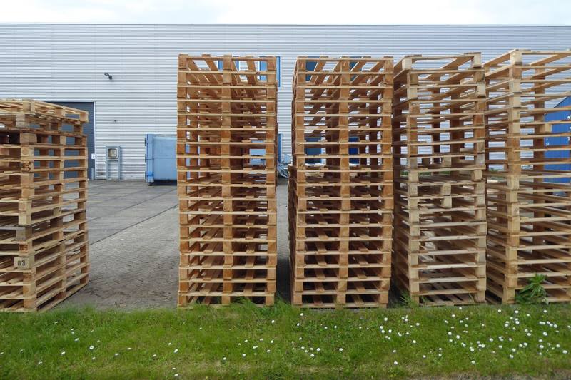 Tall stacks of empty wooden pallets on the grass outside a metal warehouse