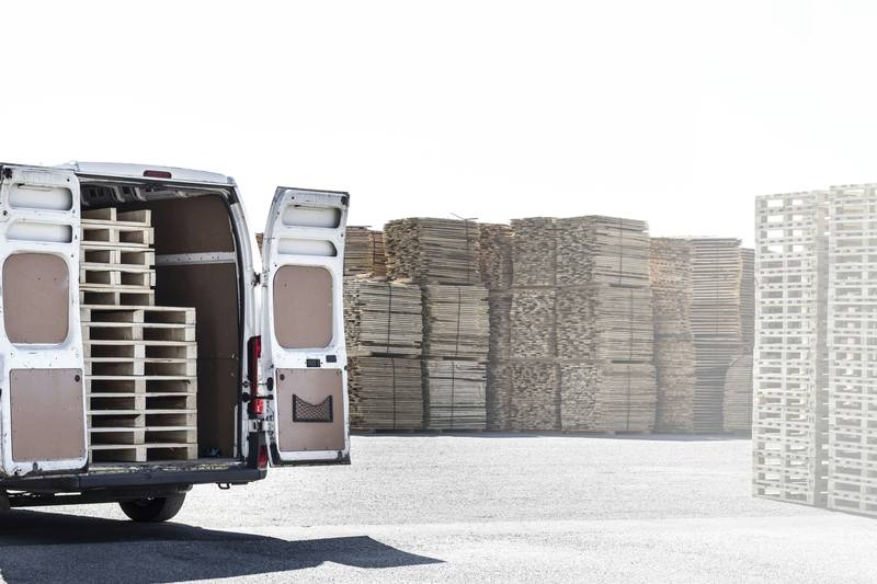 A white cargo van with its back doors open, loaded with pallets, parked in a pallet yard